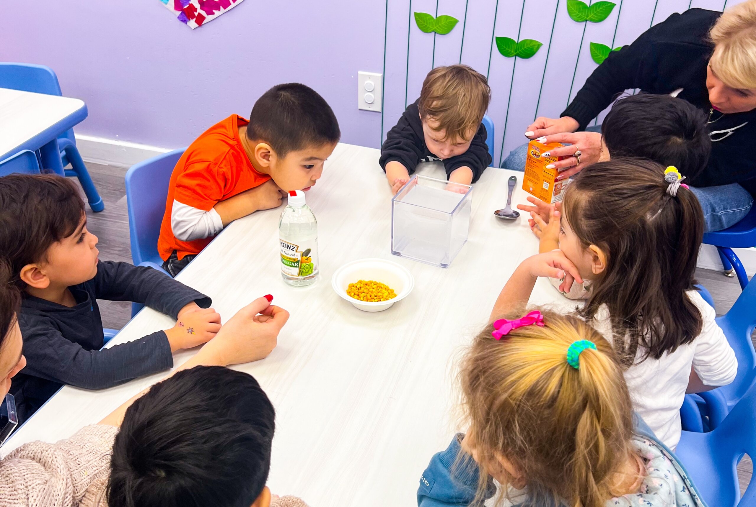 Group of young children gathered around a table, participating in a science experiment with vinegar and baking soda, supervised by an adult in a classroom with colorful decorations