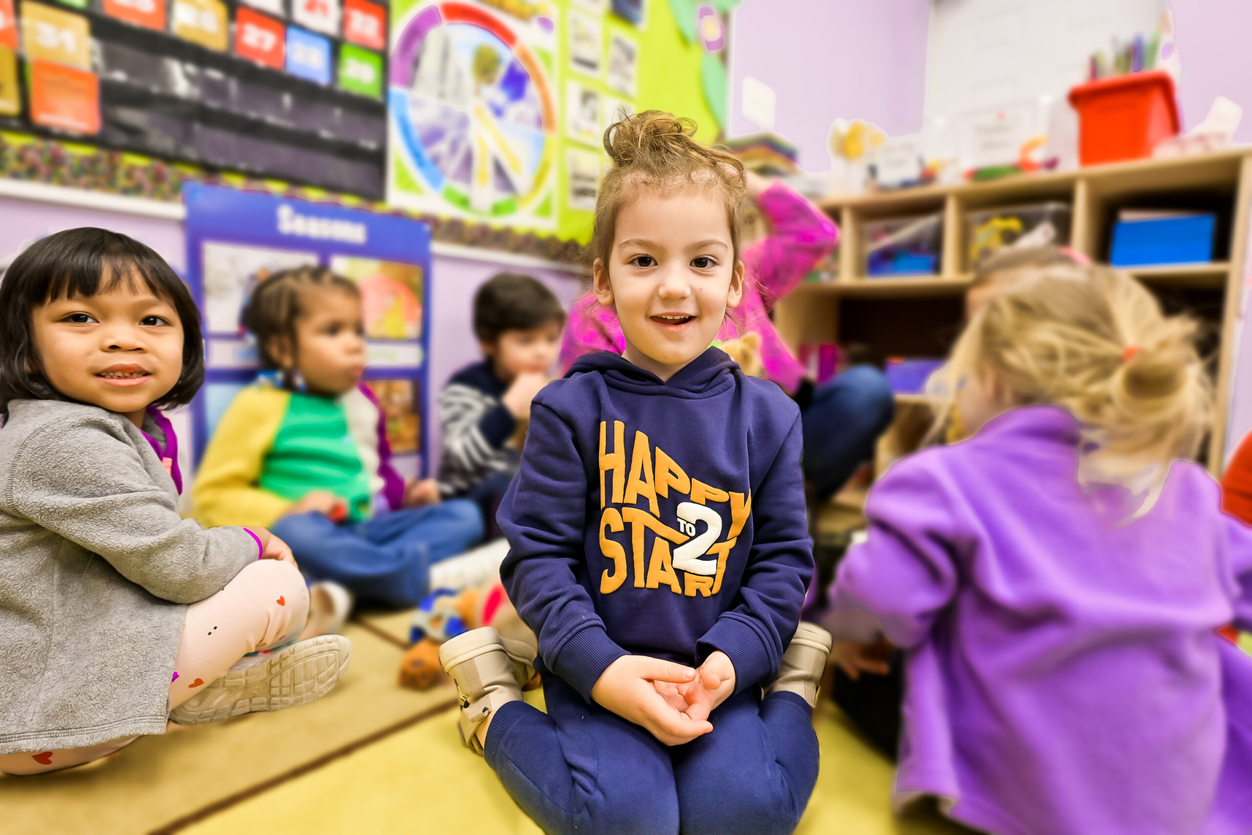 Group of children sitting on the floor in a classroom, with one child in the foreground wearing a navy blue hoodie that says 'Happy to Start,' smiling and looking at the camera