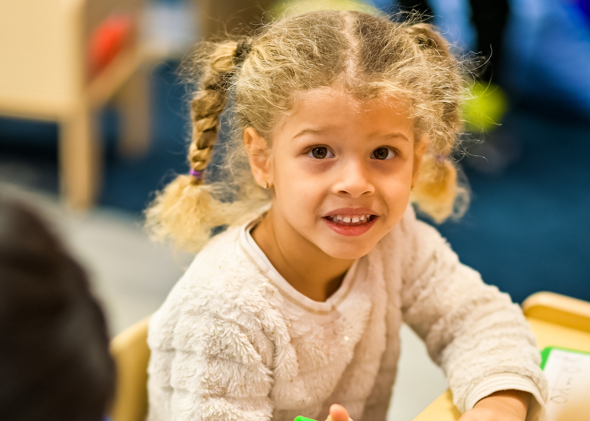 Young girl with curly pigtails, smiling and wearing a white sweater in a classroom.