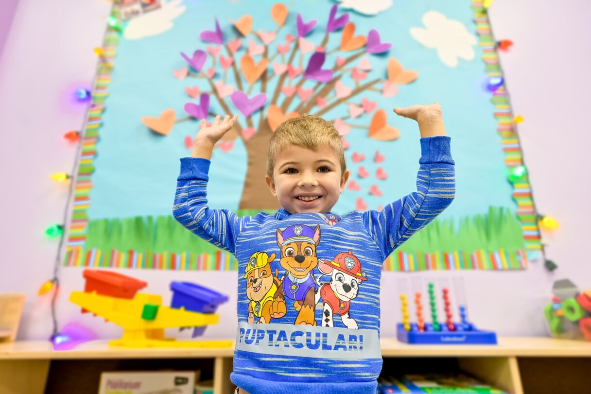 Joyful child with arms raised in a classroom decorated for St. Patrick's Day with heart-shaped tree on bulletin board