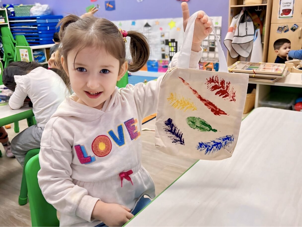 Smiling young girl displaying her colorful feather painting art project at Little Scholars Daycare.