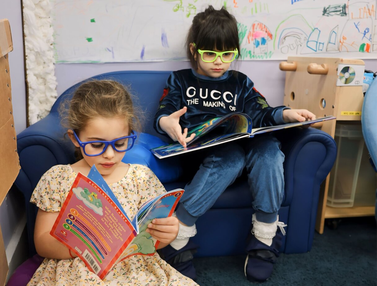 Two children with blue glasses focused on reading educational books in a colorful classroom