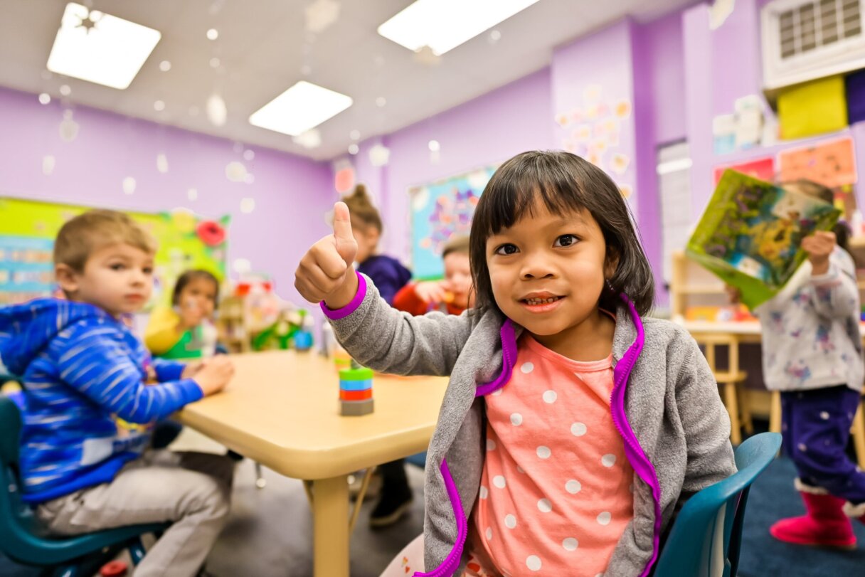 Enthusiastic preschool girl giving a thumbs up in a vibrant classroom setting