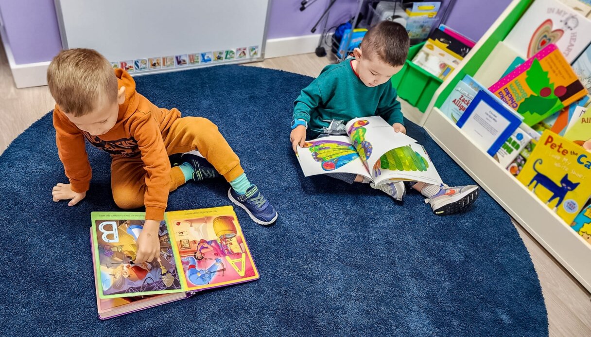 Two preschoolers reading colorful storybooks on a classroom floor, engaging in early literacy activities