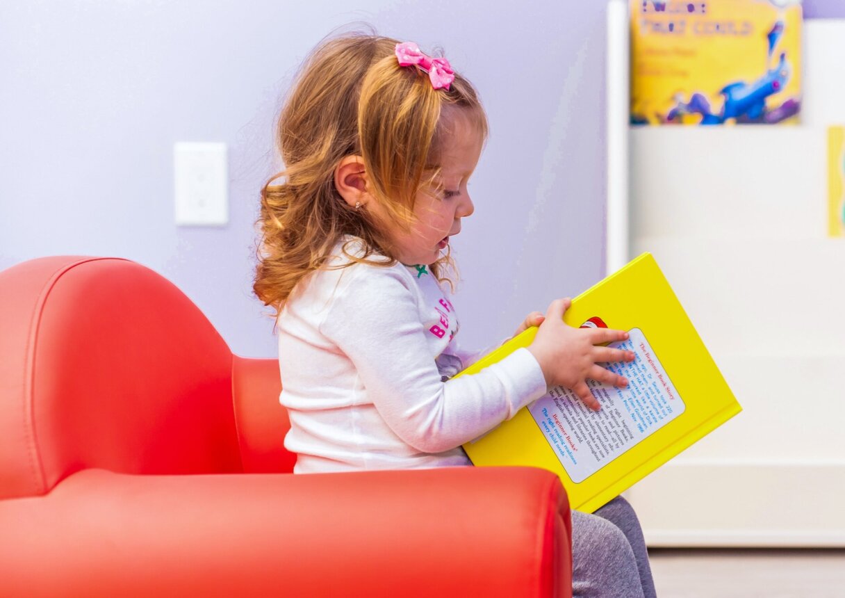 Curious toddler girl with pink hair bow reading a bright yellow book in a cozy nursery corner