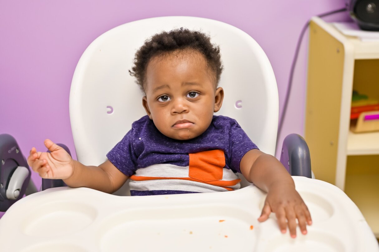 Toddler sitting thoughtfully in a high chair at daycare, adapting to mealtime after the time change.