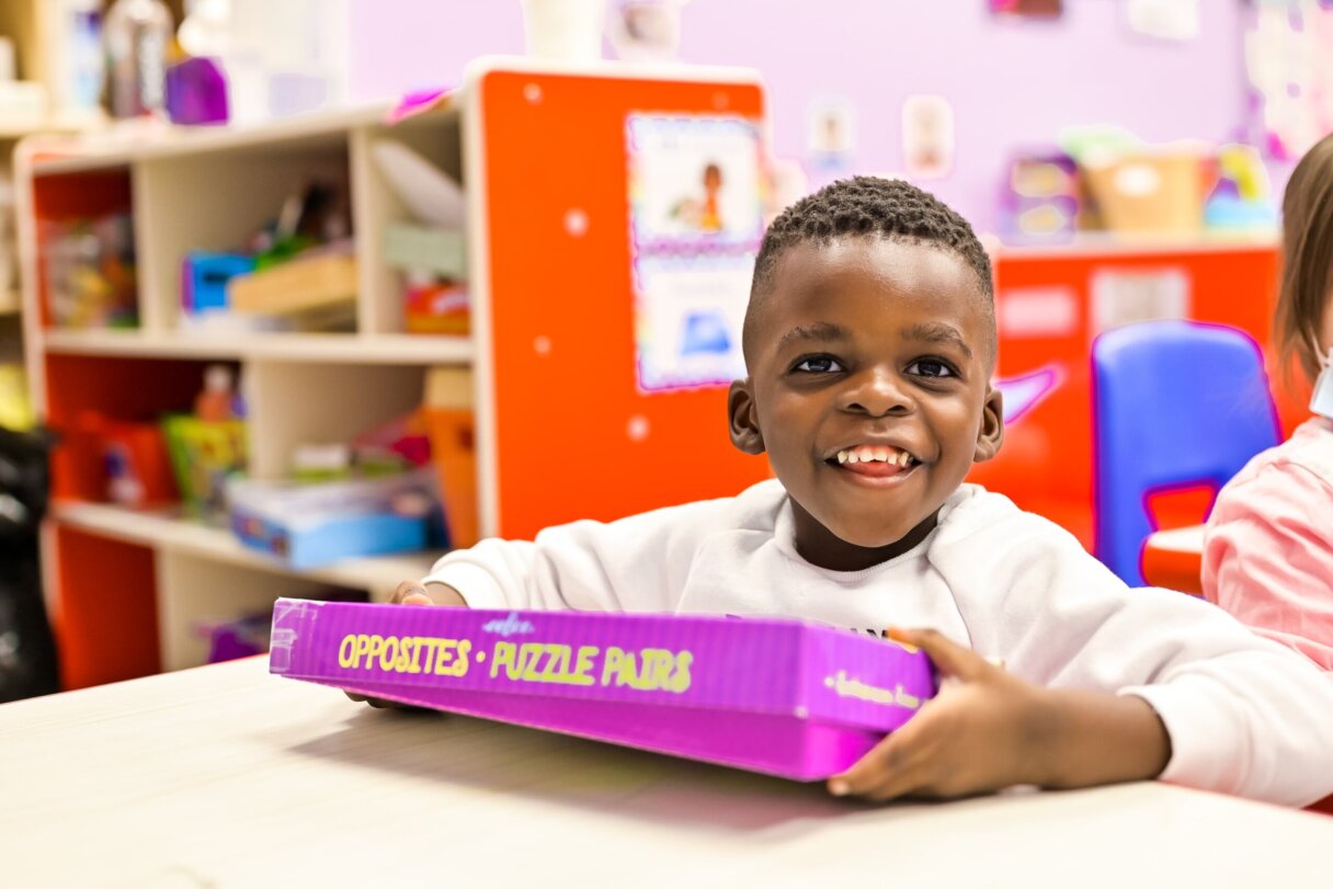 Cheerful toddler holding an 'Opposites Puzzle Pairs' box in a learning environment