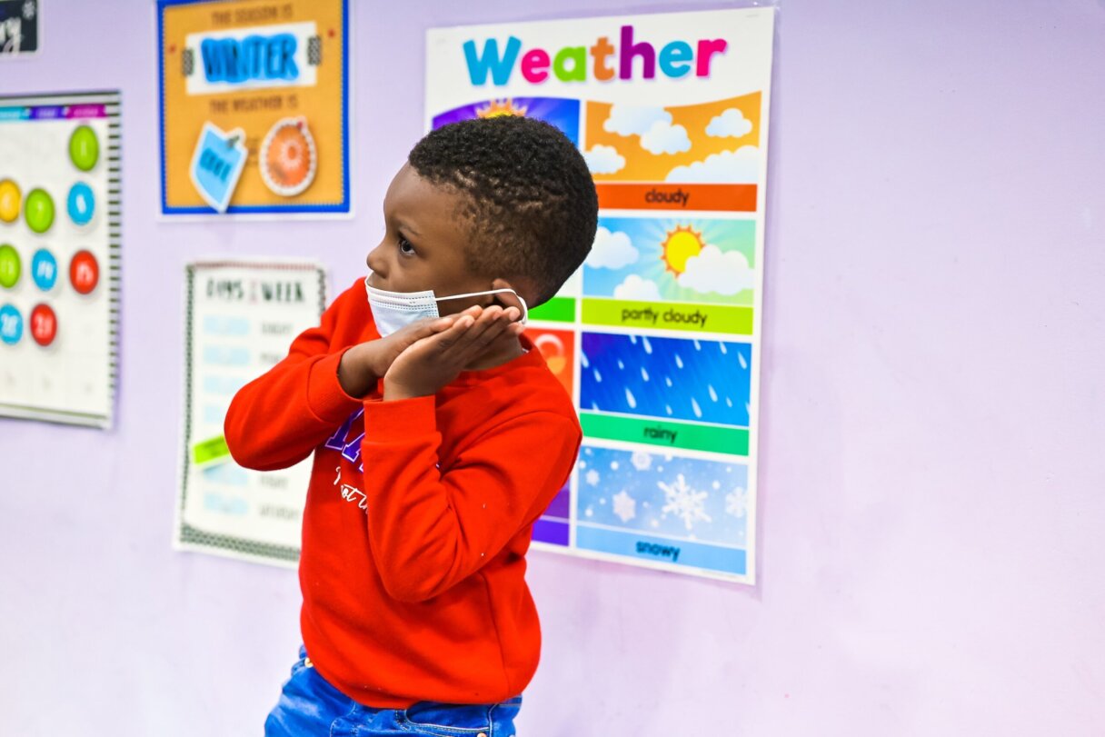Toddler gesturing in classroom with weather chart to encourage vocabulary learning