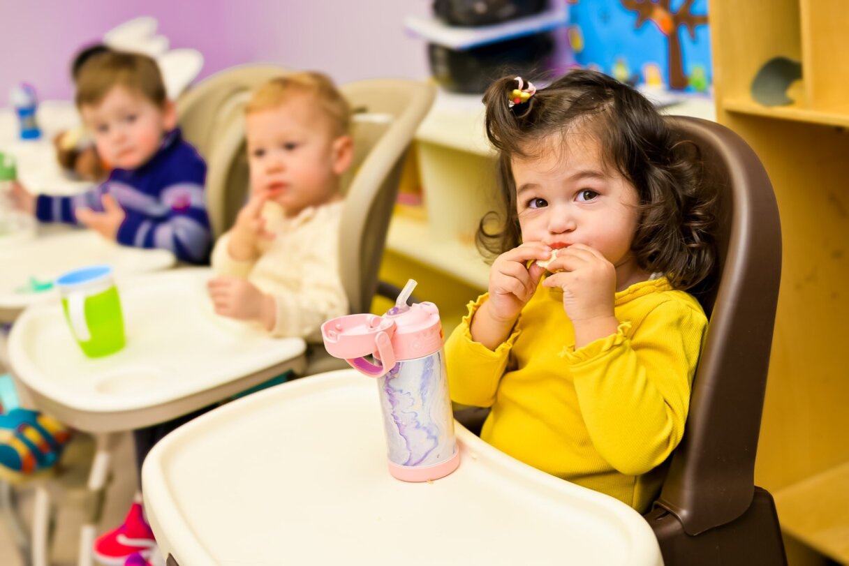 Toddler in a yellow shirt enjoying snack time with friends at preschool