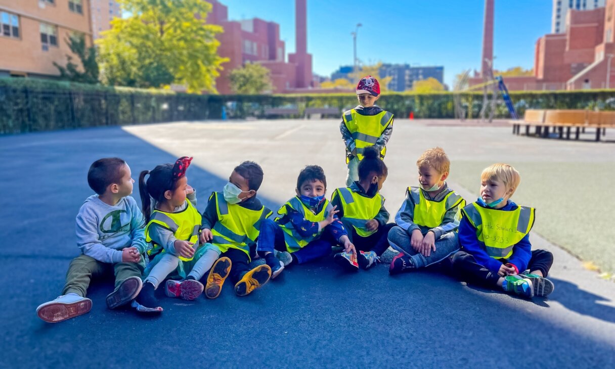 Group of children in safety vests sitting on a playground at Little Scholars Daycare.